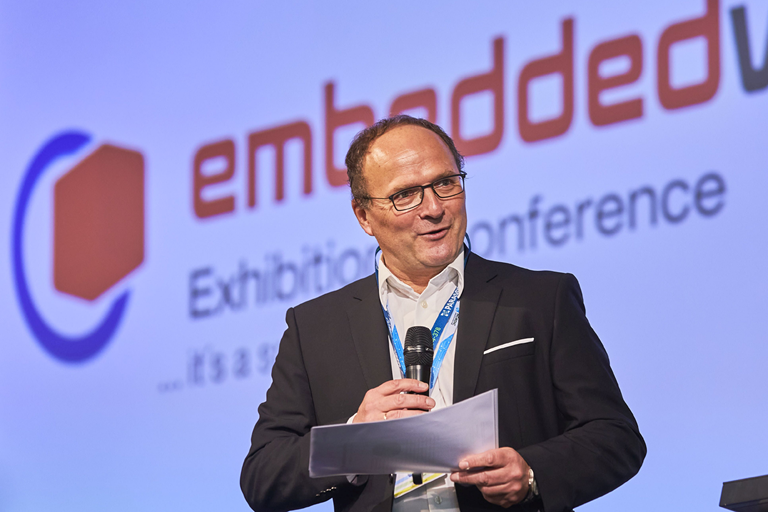 Prof. Sikora mit Mikrofon vor "embeddedworld" Hintergund