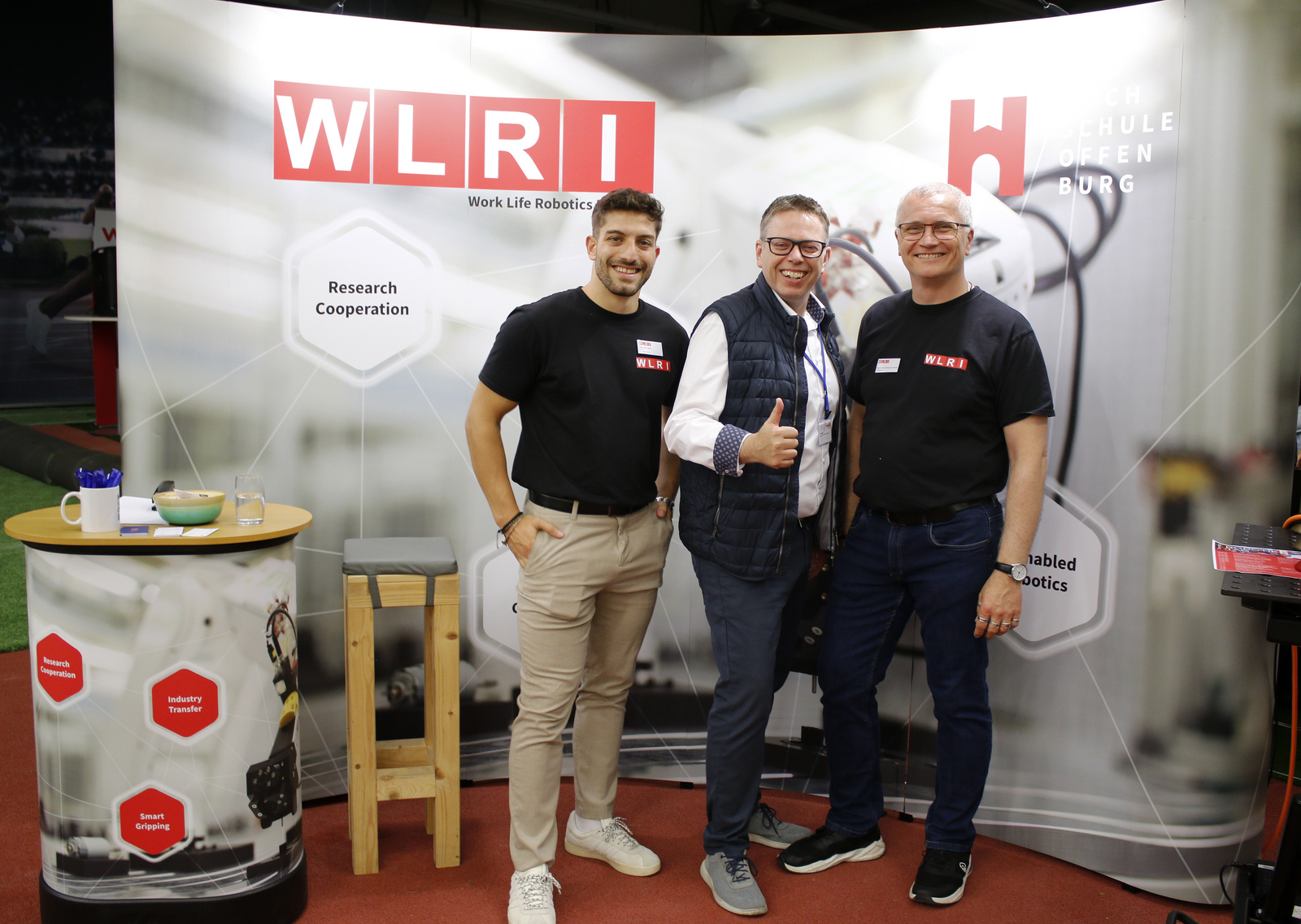 Drei Männer stehen vor eine WLRI-WAnd, der mittlere streckt den Daumen nach oben