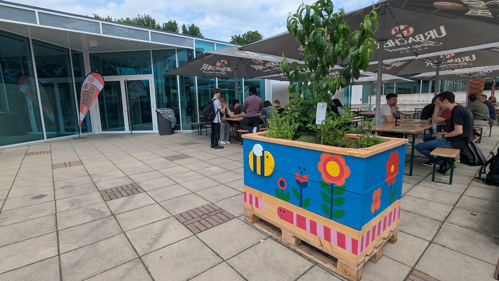 Ein Hochbeet an seinem endgültigen Standort vor der Mensa an den Tischen dahinter sitzen Leute