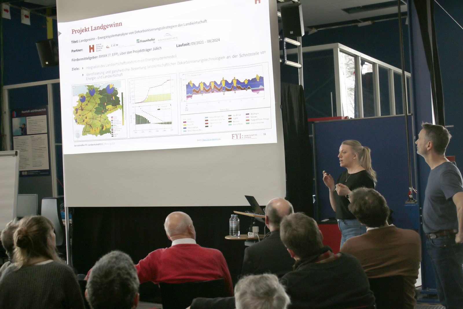 Eine Frau und ein Mann stehen recht neben einer Leinwand und erläutern den sitzenden Zuhörenden eine Präsentation