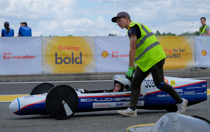 Der Schluckspecht S6, ein zigarrenförmiges blau-weißes Fahrzeug, wird mit einer Pilotin darin von einem Mann in Warnweste an den Start geschoben  