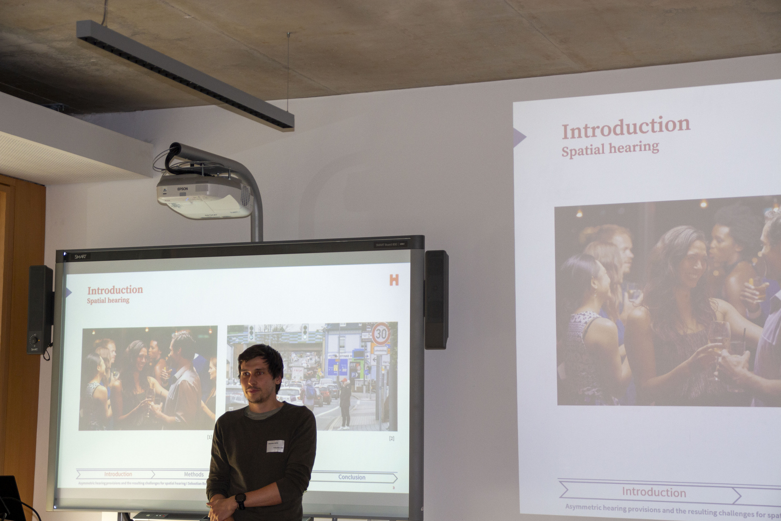 Ein junger Mann steht in einem Hörsaal vor einem Bildschirm auf dem eine Präsentation zu sehen ist