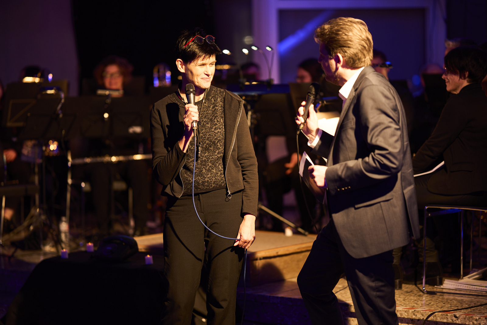 Eine Frau und ein Mann stehen vor dem sitzenden Orchester und unterhalten sich