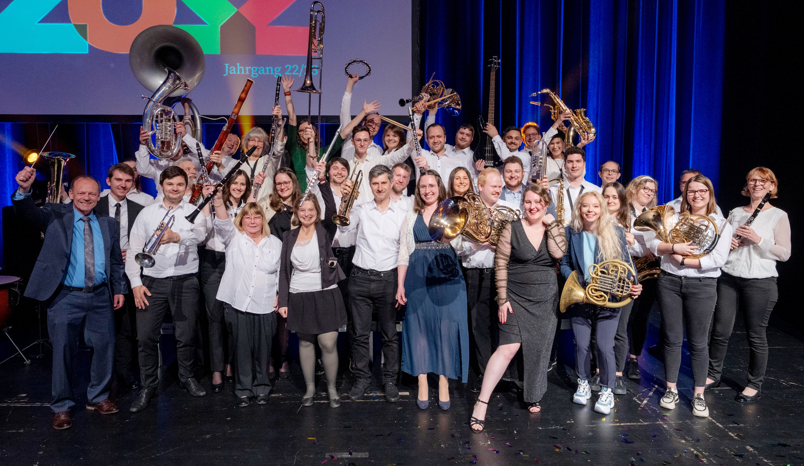 Gruppenfoto des stehenden und jubelnd die Instrumente in die Höhe reckenden Orchesters auf einer Bühne 