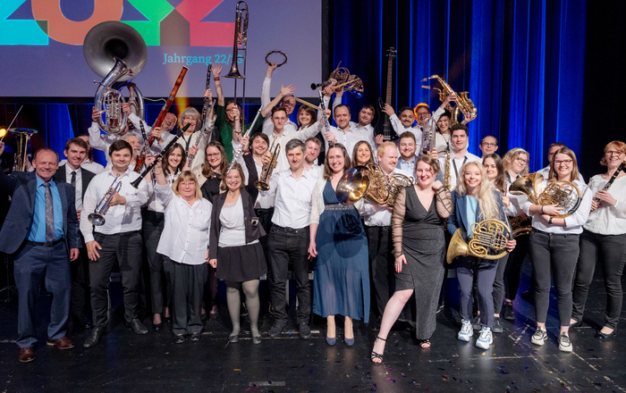 Gruppenfoto des stehenden und jubelnd die Instrumente in die Höhe reckenden Orchesters auf einer Bühne 