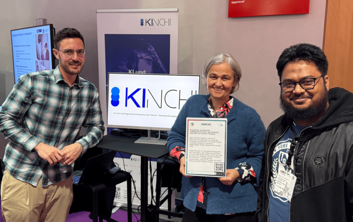 Ein Gruppenfoto von drei lächelnden Personen vor einem Messestand des Projekts KINCHI auf der Zukunft Handwerk 2025.