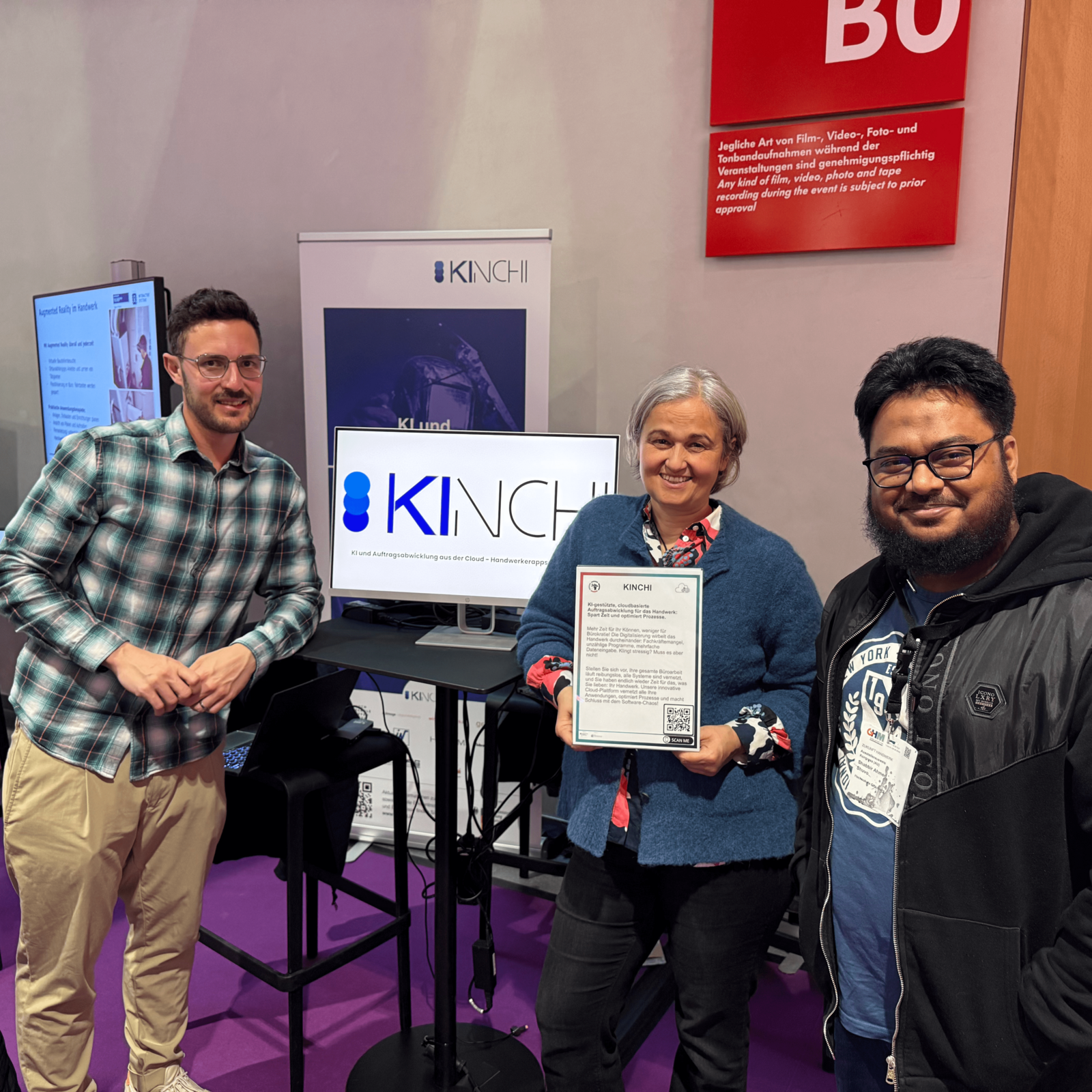 Ein Gruppenfoto von drei lächelnden Personen vor einem Messestand des Projekts KINCHI auf der Zukunft Handwerk 2025.