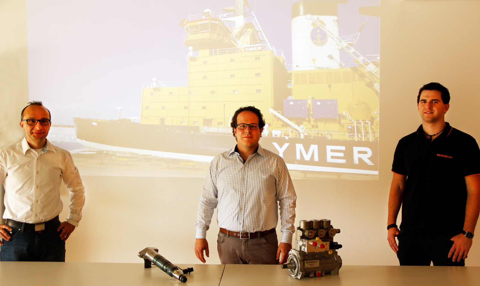 Die HEINZMANN-Entwicklungsingenieure und ehemaligen Maschinenbau-Studenten der Hochschule Offenburg Simon Serrer (Mitte) und Andreas Ruf (rechts) mit ihrem früheren Professor Claus Fleig (links) vor einem per Beamer an die Wand geworfenen Bild eines Containerschiffs.