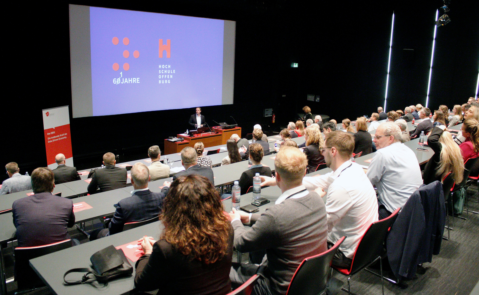 Ein Mann steht vor einer Leinwand an einem Rednerpult und spricht zu den im Hörsaal Sitzenden