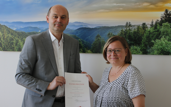 Mann übergibt Urkunde an Frau. Beide stehen vor einem Schwarzwaldfoto 