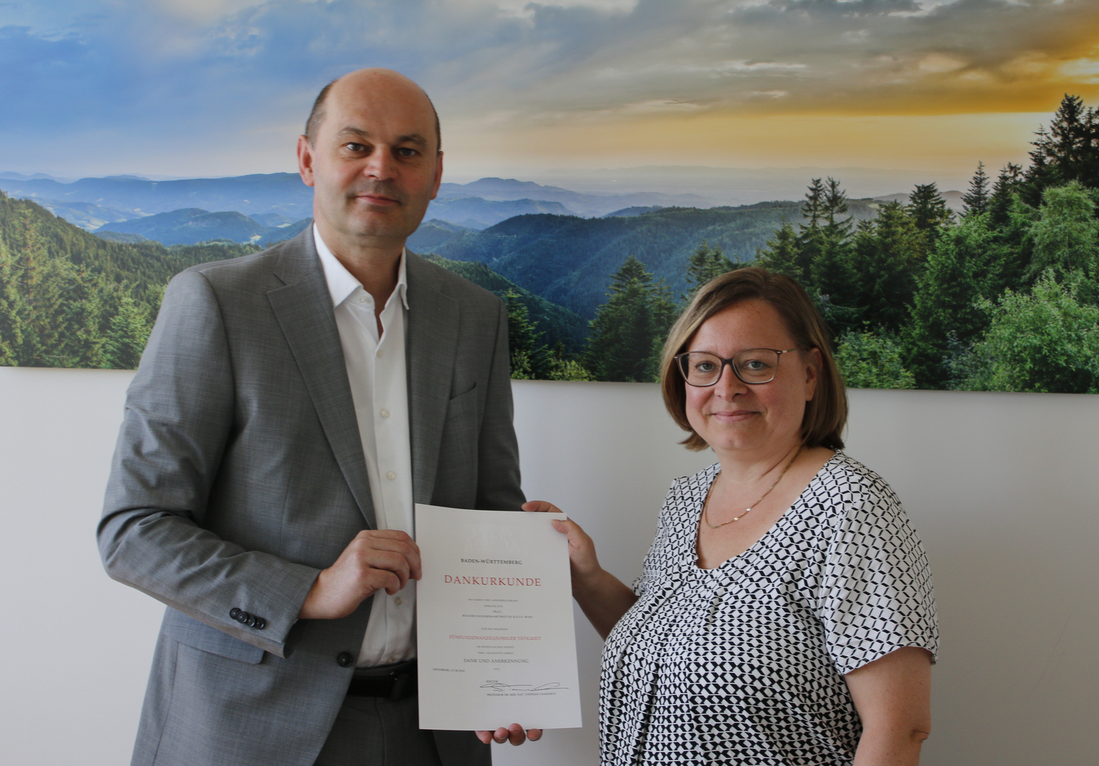 Mann übergibt Urkunde an Frau. Beide stehen vor einem Schwarzwaldfoto 