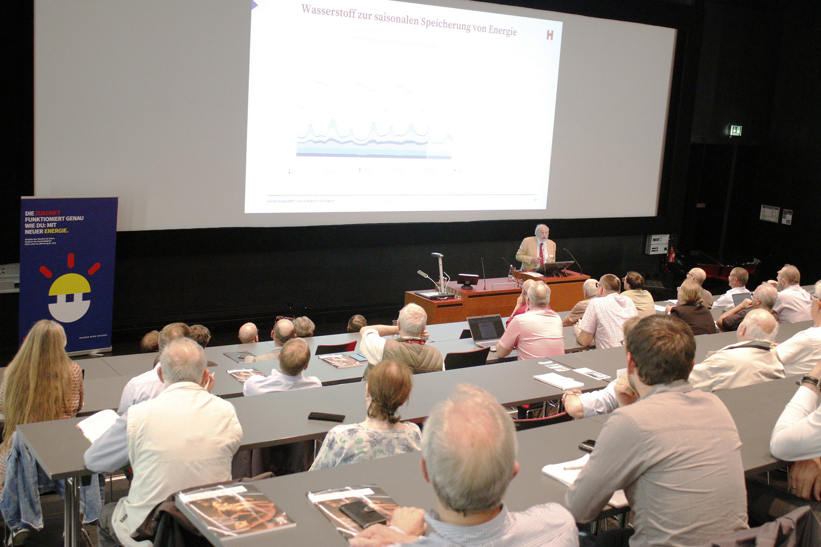 Mann steht vor den sitzenden Zuhörenden im Hörsaal, im Hintergrund ist eine Leinwand 