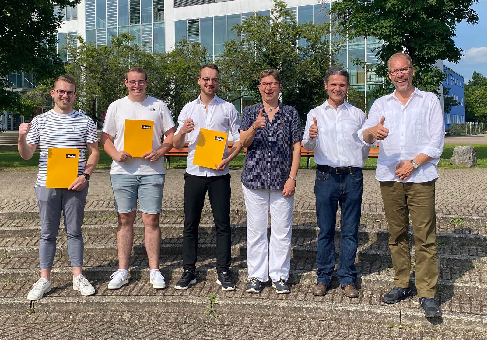 Der Preisstifter, die Preisträger und die Dekan*innen der beiden Fakultäten stehen auf dem Campus und machen das Daumen hoch-Zeichen  