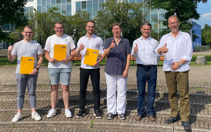 Der Preisstifter, die Preisträger und die Dekan*innen der beiden Fakultäten stehen auf dem Campus und machen das Daumen hoch-Zeichen  