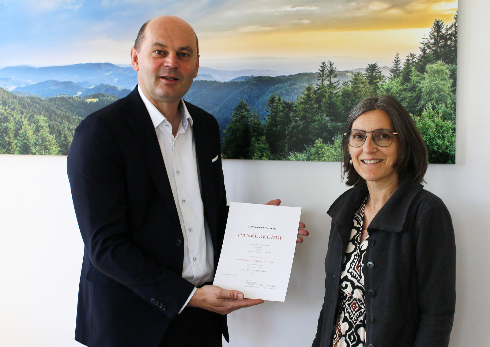 Ein Mann und eine Frau stehen vor einem Schwarzwaldbild, der Mann hält eine Urkunde in den Händen