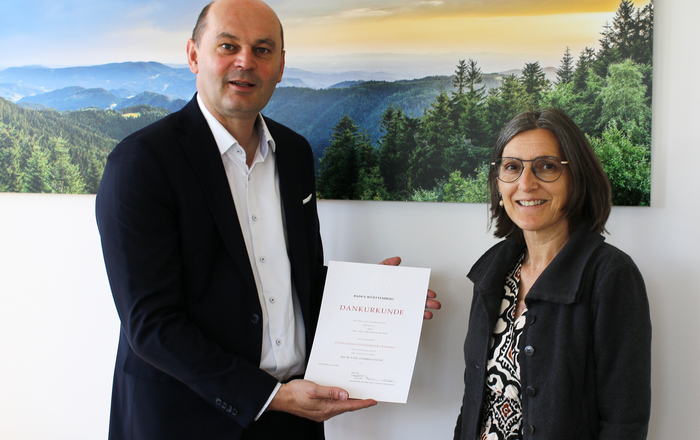 Ein Mann und eine Frau stehen vor einem Schwarzwaldbild, der Mann hält eine Urkunde in den Händen