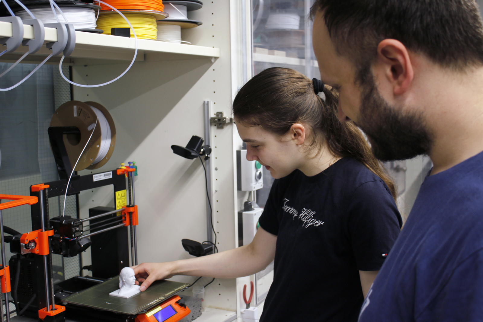 Ein Mädchen und ein Mann stehen an einem 3D-Drucker in dem ein kleiner Kopf steht