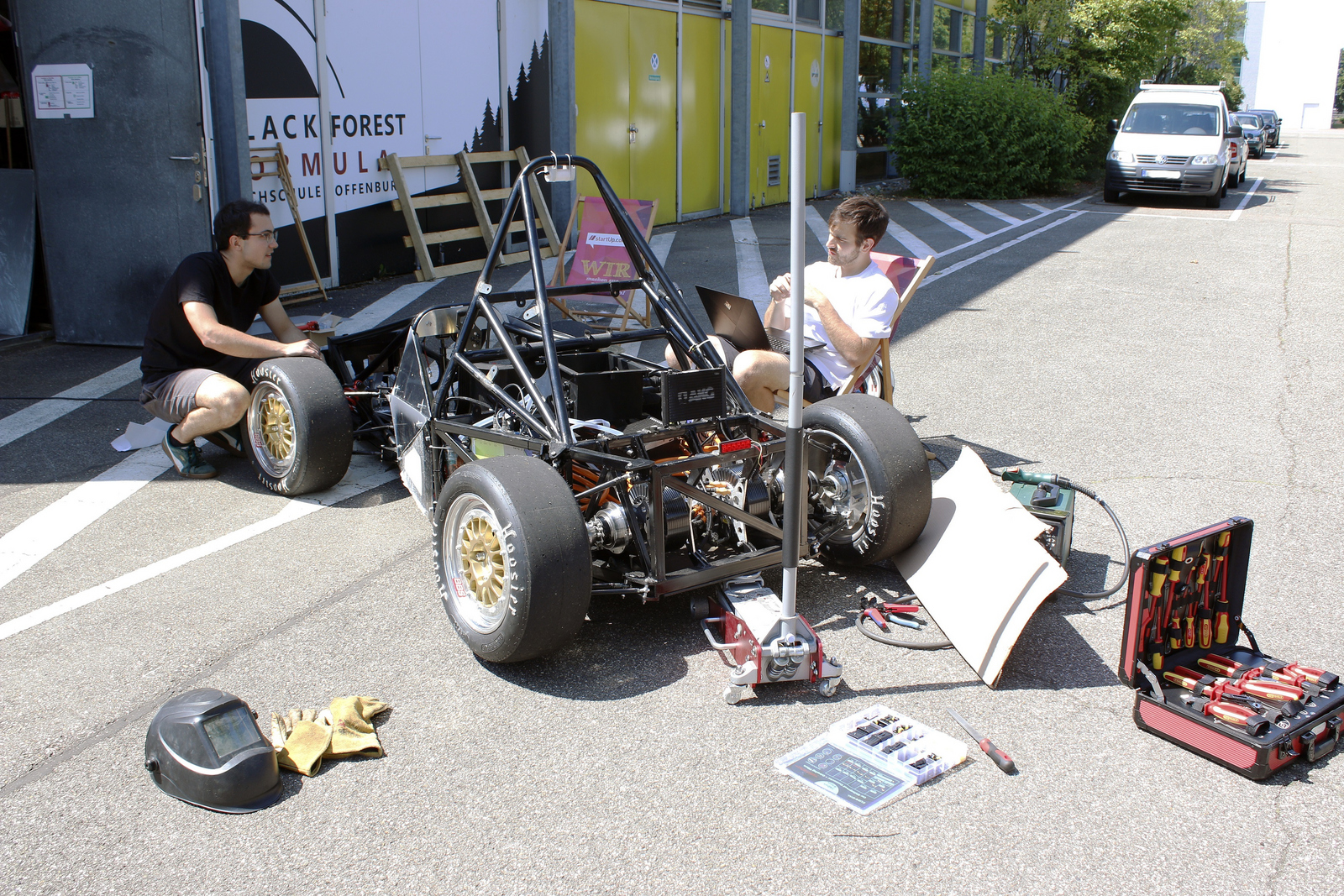 Blick von hinten auf den Rennwagen: Ein junger Mann kniet neben dem linken Vorderreifen des Rennwagens und schraubt daran, ein anderer sitzt rechts neben dem Rennwagen in einem Liegestuhl und hat ein Laptop auf dem Schoß überall liegt Werkzeug herum 