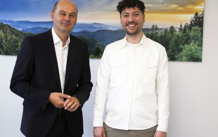 Zwei Männer stehen vor einem Schwarzwaldfoto und schauen in die Kameraa