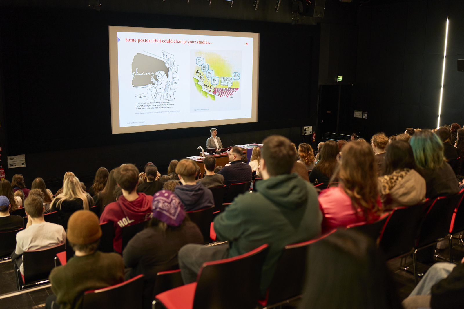 Eine Frau steht vor sitzenden jungen Menschen in einem Hörsaal hinter ihr sind zwei Bilder auf einer Leinwand zu sehen