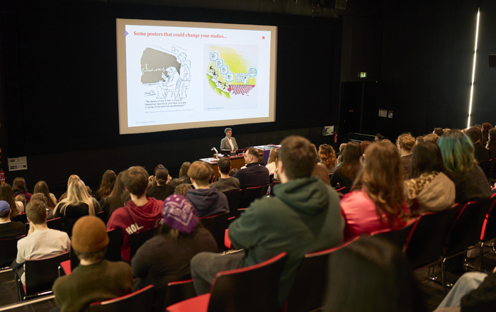 Eine Frau steht vor sitzenden jungen Menschen in einem Hörsaal hinter ihr sind zwei Bilder auf einer Leinwand zu sehen