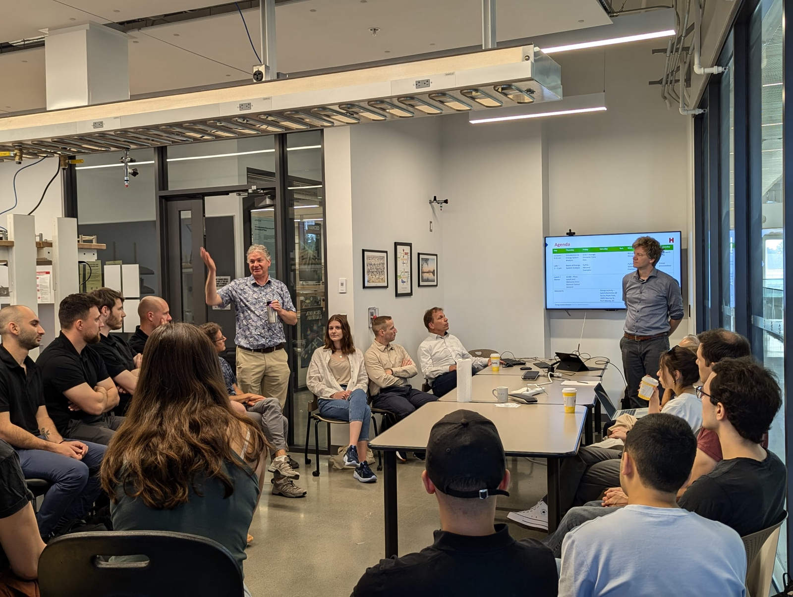 Zwei Männer stehen vor jungen Leuten und erläutern eine Agenda auf einem Bildschirm