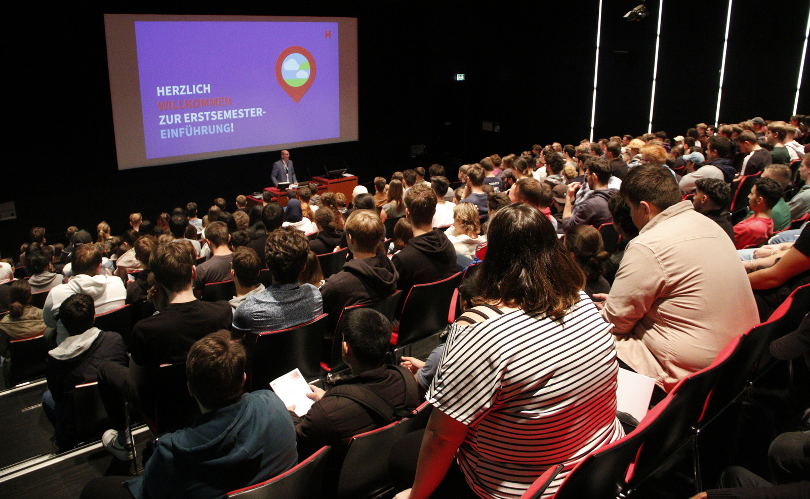 Der Rektor steht vor den Erstsemestern im vollbesetzten Winfried-Lieber-Saal am Rednerpult. Hinter ihm ist an eine Leinwand eine Präsentation zu erkennen. im  