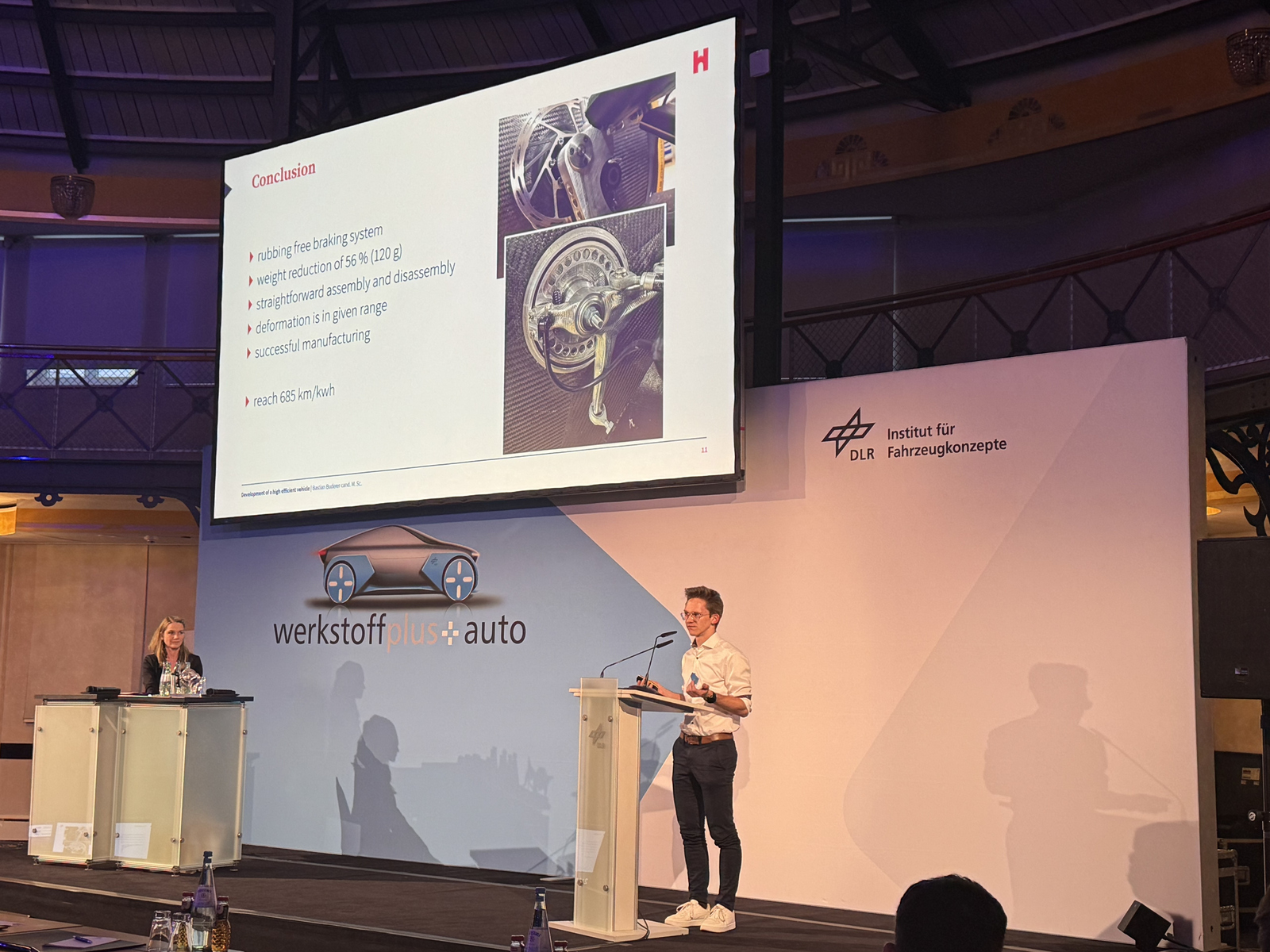 Ein junger Mann steht am Rednerpult und erläutert eine Prasentaion auf einem großen Bildschirm