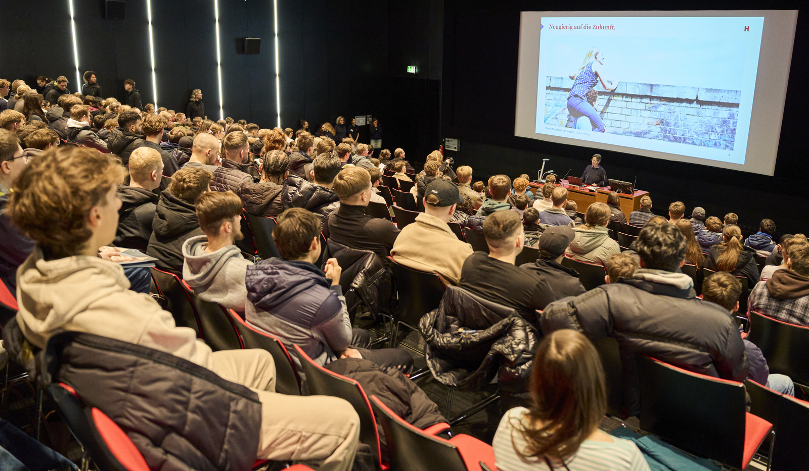 Blick über in einem Hörsaal sitzende Schülerinnen und Schüler hin zum Rednerpult und der Leinwand dahinter