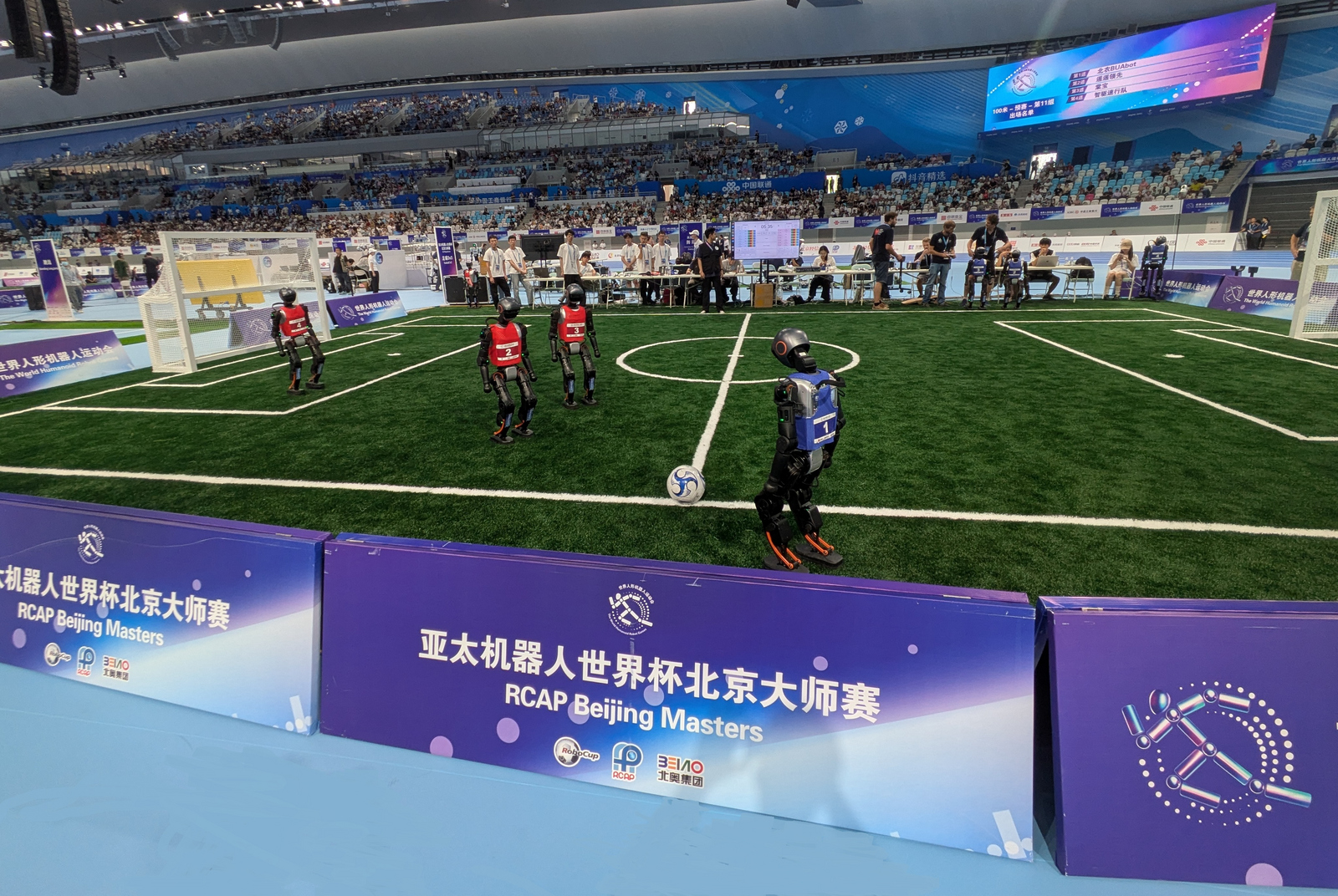 Auf einem Fuballfeld in einer Halle spielen drei rotgkleidete Roboter gegen drei blaugekleidete, von denen einer gerade eine Freistoß an der Seitenlinie ausführt. Am Spielfeldrand stehen Menschen und auf den Tribünen sitzen zahlreiche Zuschauende.