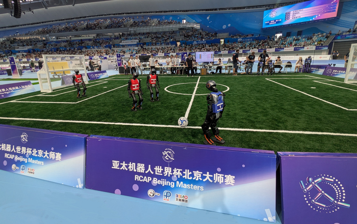 Auf einem Fuballfeld in einer Halle spielen drei rotgkleidete Roboter gegen drei blaugekleidete, von denen einer gerade eine Freistoß an der Seitenlinie ausführt. Am Spielfeldrand stehen Menschen und auf den Tribünen sitzen zahlreiche Zuschauende.