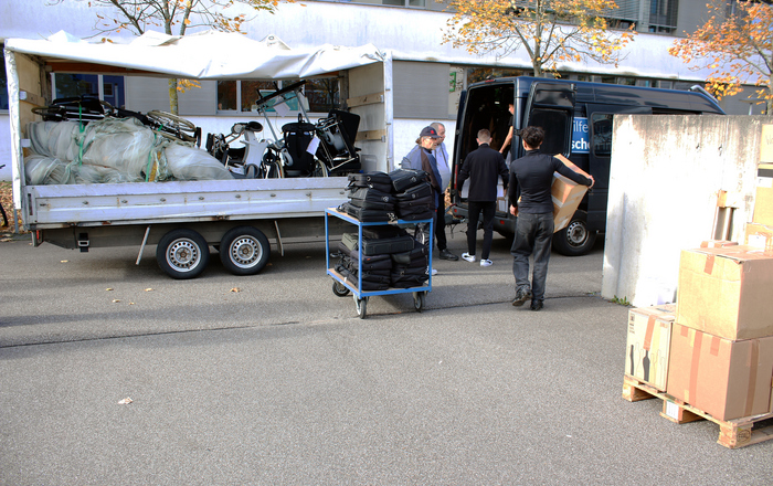 Mehrere Männer packen Kisten und Kaptops in einen Kastenwagen mit Anhänger auf dem Rollstühle und Netze aus dem Obst- und Weinbau liegen