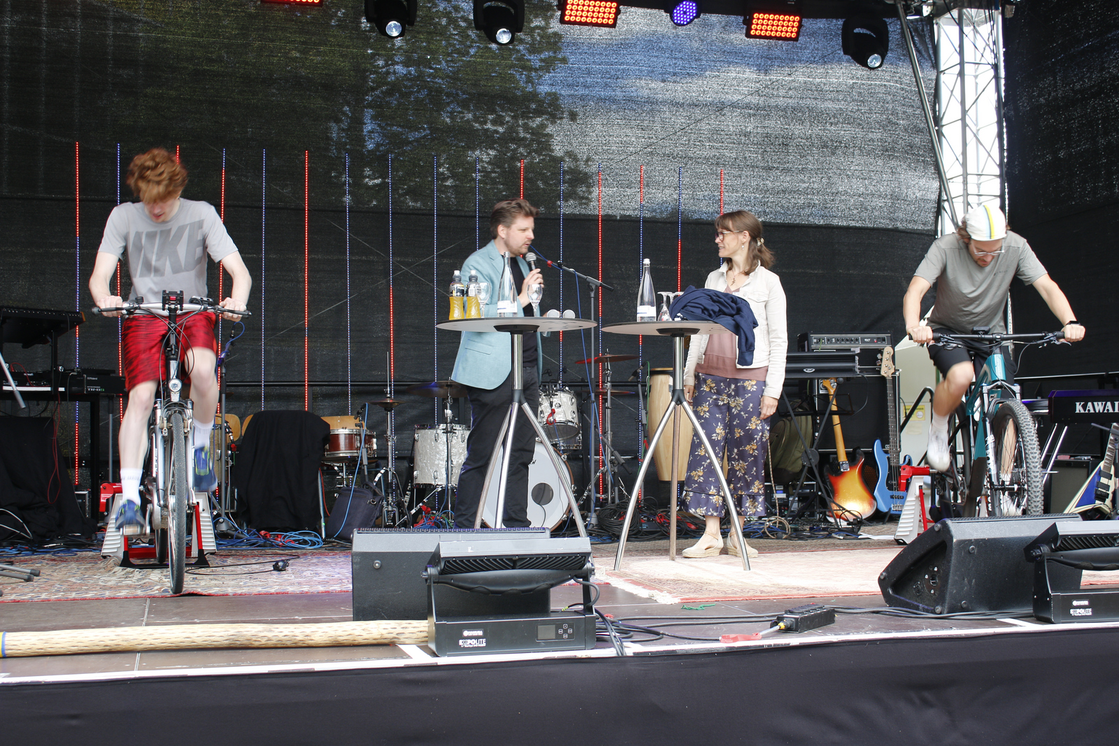 Zwei Männer strampeln auf den beiden Rädern, zwischen ihnen stehen ein MAnn und eine Frau und reden.