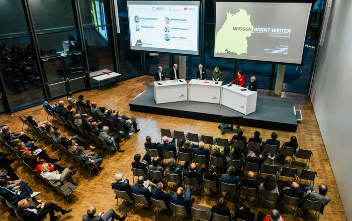 Mehrere Menschen stehen auf einer Bühne im Halbkreis an Rednerpulten hinter ihne zwei große Bidlschirme mit Präsentationen und vor ihnen sitzende Zuhörende