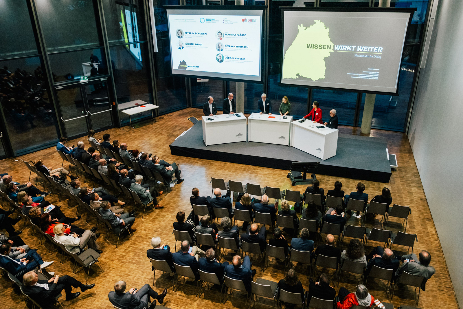 Mehrere Menschen stehen auf einer Bühne im Halbkreis an Rednerpulten hinter ihne zwei große Bidlschirme mit Präsentationen und vor ihnen sitzende Zuhörende