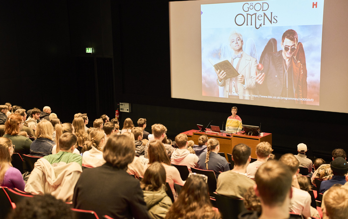 Eine Frau steht vor sitzenden jungen Menschen in einem Hörsaal hinter ihr auf eine Leinwand ist das Titelbild der Serie Good Omens zu sehen