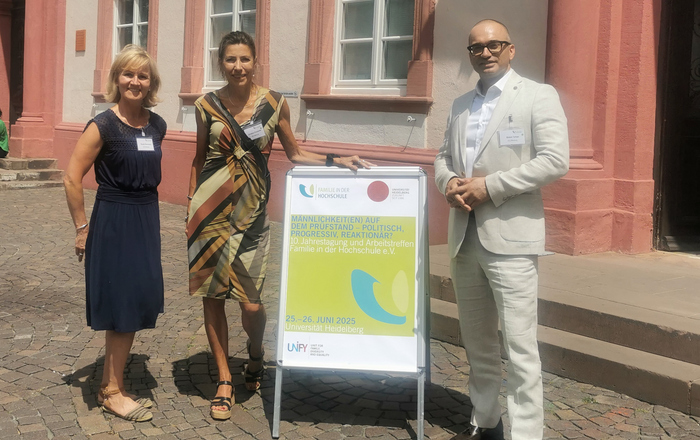 Zwei Frauen in Kleidern stehen links, ein Mann im Anzug rechts von einem Aufsteller vor der Uni Heidelberg.