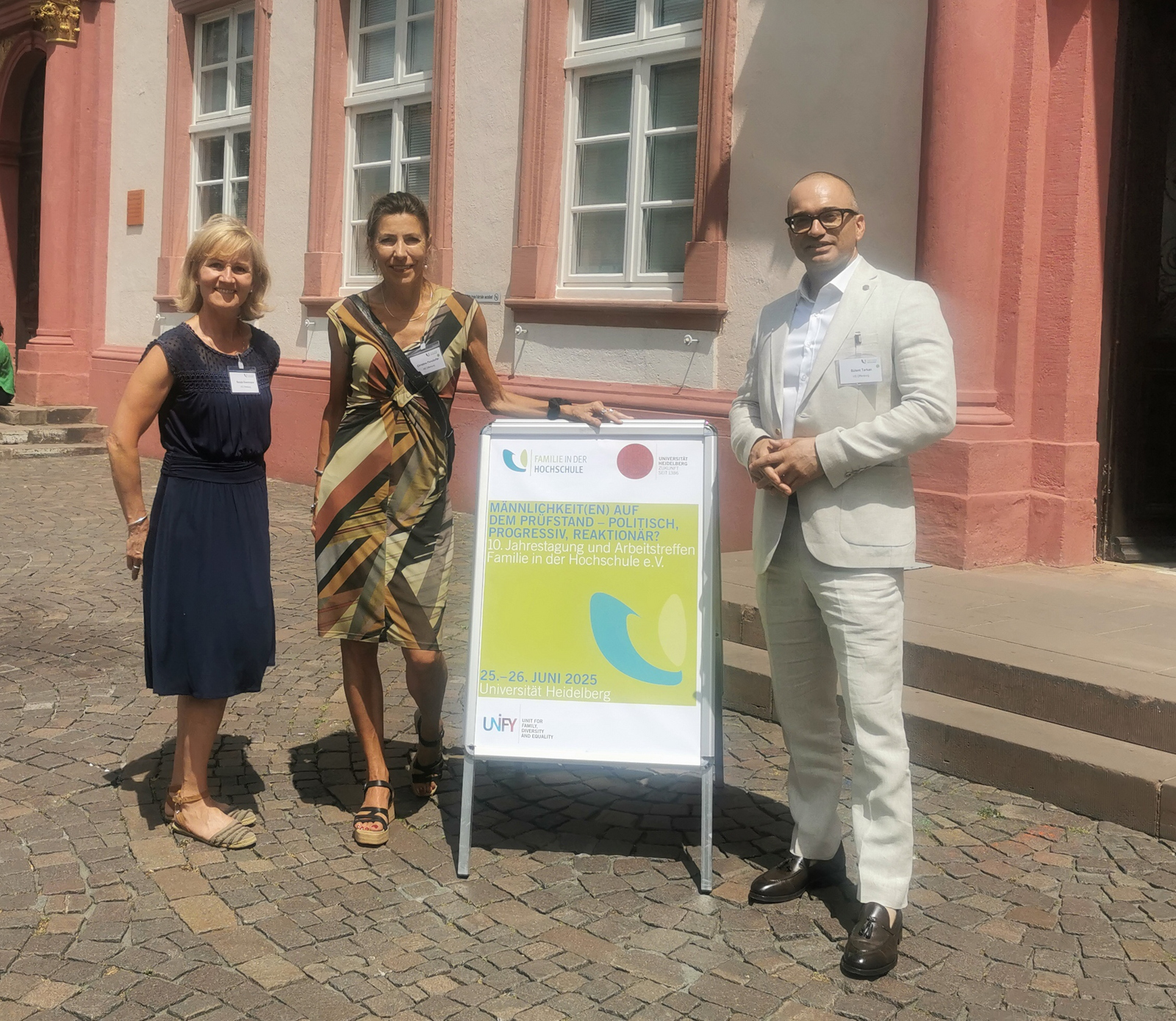 Zwei Frauen in Kleidern stehen links, ein Mann im Anzug rechts von einem Aufsteller vor der Uni Heidelberg.