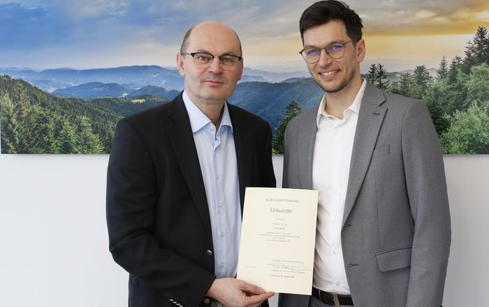 Zwei Männer im Anzug stehen vor eine Schwarzwaldfoto und halten eine Urkunde zwischen sich
