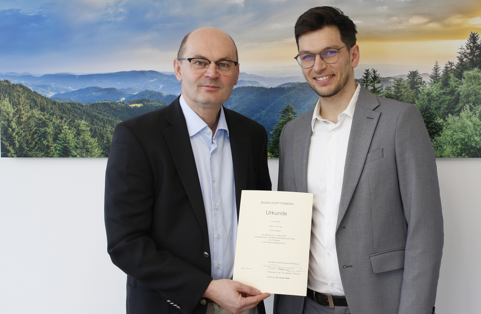 Zwei Männer im Anzug stehen vor eine Schwarzwaldfoto und halten eine Urkunde zwischen sich