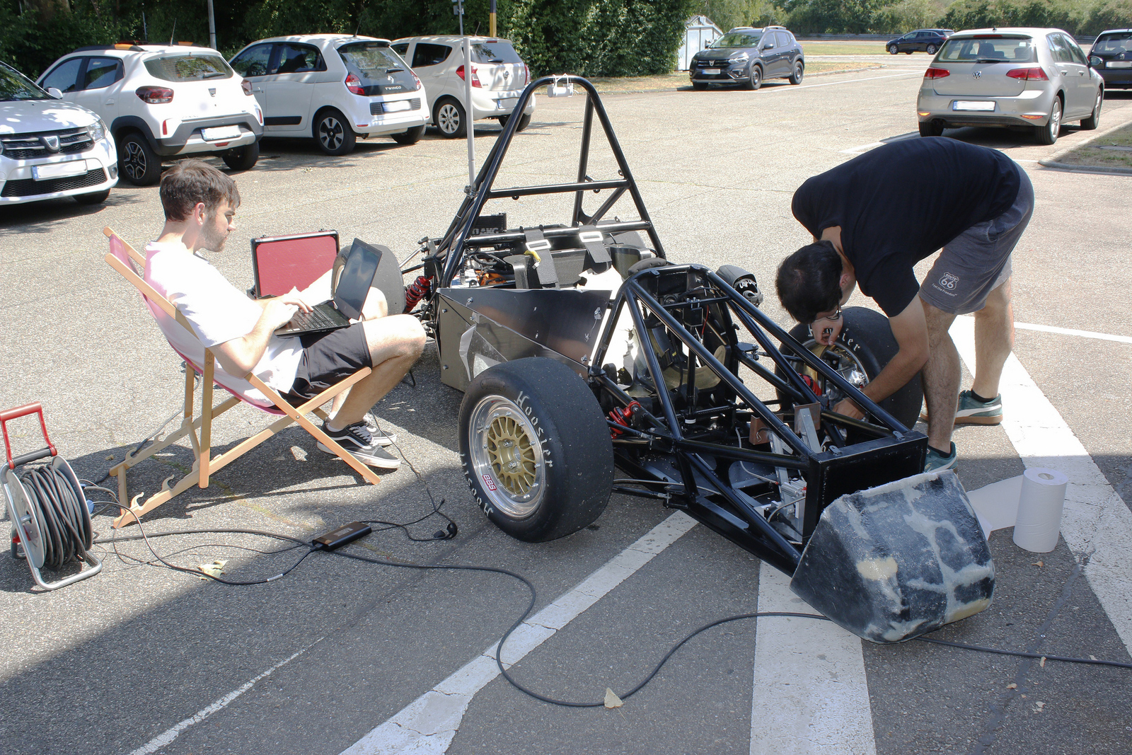 Blick von ron auf den Rennwagen: Ein junger Mann beugt sich über den linken Vorderreifen des Rennwagens und schraubt daran, ein anderer sitzt rechts neben dem Rennwagen in einem Liegestuhl und hat ein Laptop auf dem Schoß überall liegt Werkzeug herum 