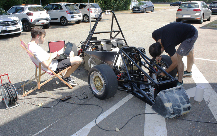 Blick von ron auf den Rennwagen: Ein junger Mann beugt sich über den linken Vorderreifen des Rennwagens und schraubt daran, ein anderer sitzt rechts neben dem Rennwagen in einem Liegestuhl und hat ein Laptop auf dem Schoß überall liegt Werkzeug herum 