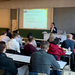 In einem Vorlesungssaal sitzen ganz viele Menschen vorne, vor einer Leinwand auf der Informationen zu lesen sind steht der Prodekan