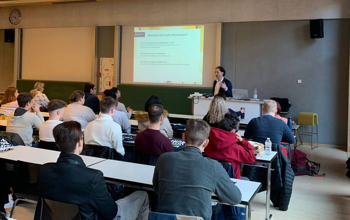 In einem Vorlesungssaal sitzen ganz viele Menschen vorne, vor einer Leinwand auf der Informationen zu lesen sind steht der Prodekan