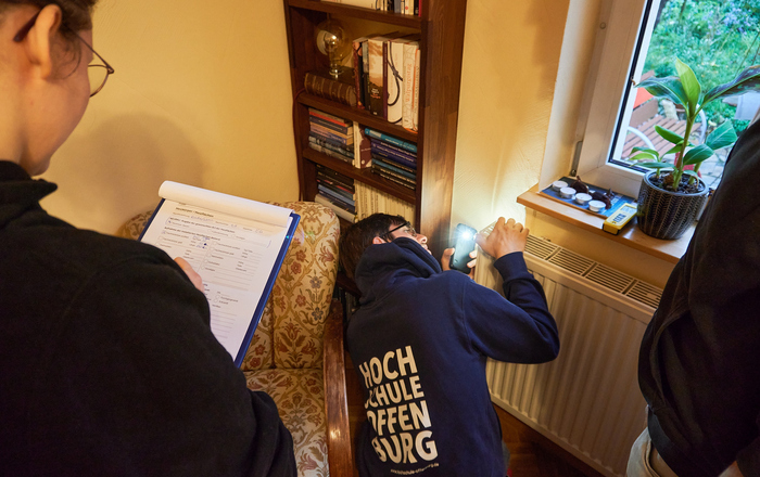 Ein Student beugt sich über einen Heizkörper und misst etwas, ein anderer schreibt die Daten auf.