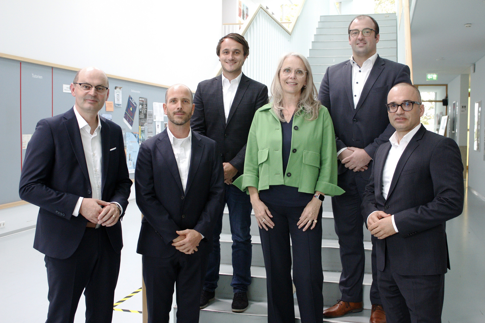 Gruppefoto von fünf Männern in dunklen Anzügen und einer Frau in grüner Kostümjacke stehen auf beziehungsweise am Fuß einer Treppe