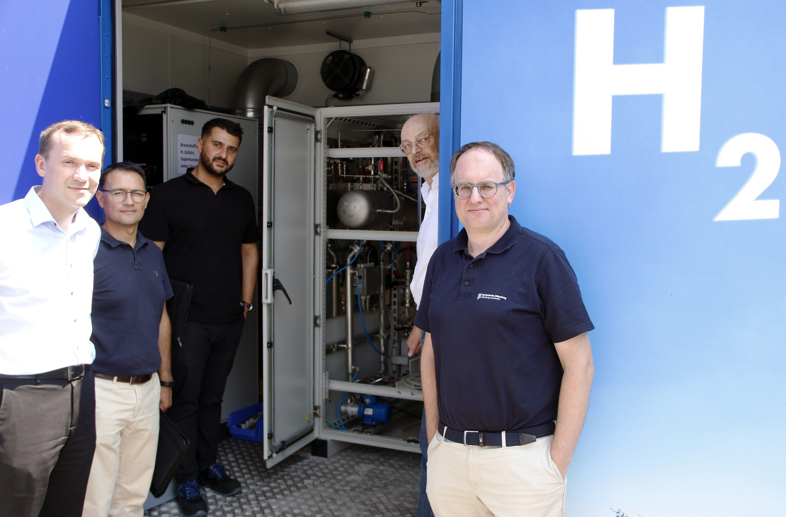 Die Herren stehen an und in der Tür des Containers in dem der Elektrolyseur untergebracht ist. Auf dem Container steht H2