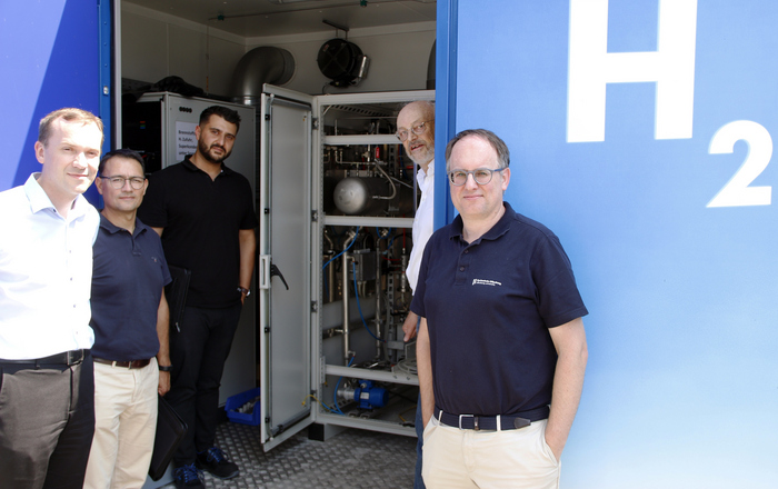 Die Herren stehen an und in der Tür des Containers in dem der Elektrolyseur untergebracht ist. Auf dem Container steht H2