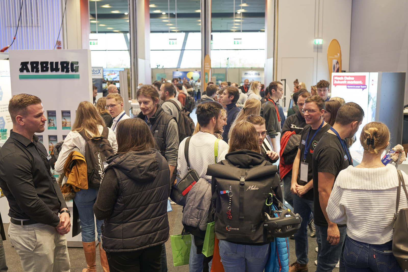 Blick vom großen Saal ins Foyer mit vielen Messeständen die umringt sind von Studierenden