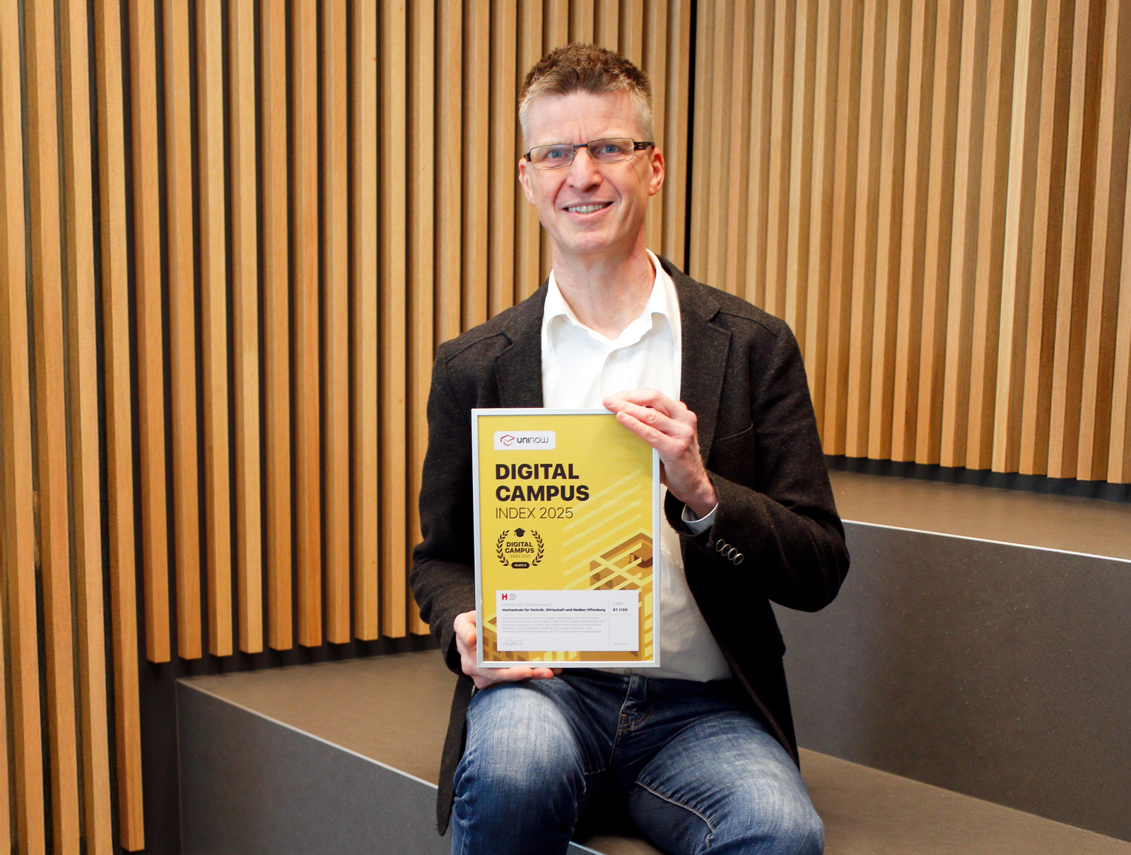 Ein Mann sitzt vor einer Wand aus Holzlamellen auf einer Treppe und präsentiert die gerahmte Urkunde 
